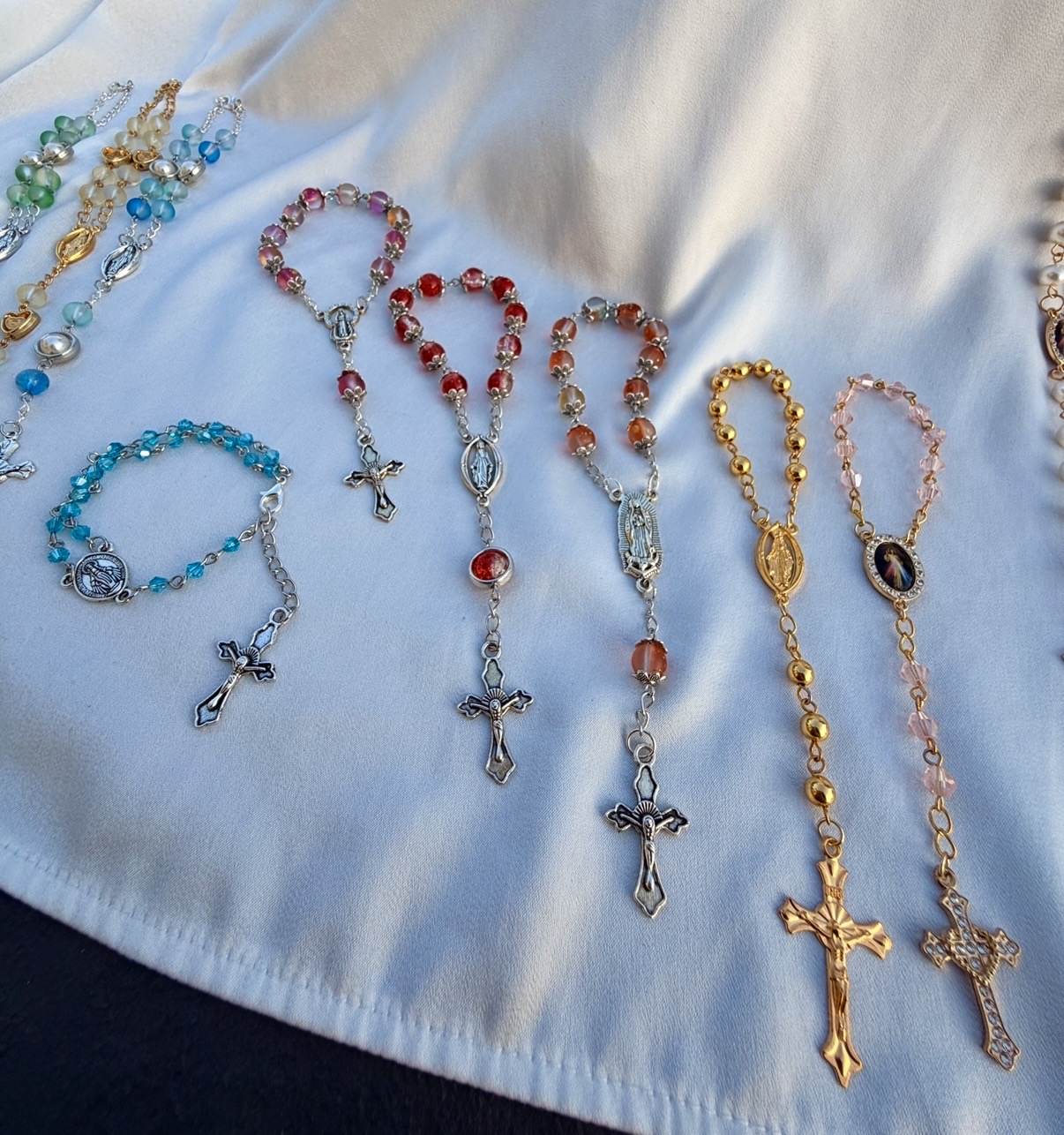 Pocket rosaries and rosary bracelets in pink, red, amber, gold and blush laid on white satin